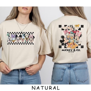 May include: A natural-colored t-shirt featuring a graphic of Disney characters including Mickey, Minnie, Donald, Daisy, Goofy, and Pluto. The front shows the characters in a row over a black and white checkerboard pattern. The back features the same characters grouped together above the text Mickey & Co. Est 1928.
