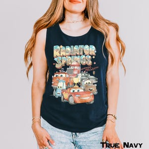 Puede incluir: Camiseta sin mangas azul marino con un gr&aacute;fico colorido de personajes de la pel&iacute;cula Cars, incluido Rayo McQueen. El texto "Radiator Springs" se muestra encima de los coches en una fuente retro. La camiseta sin mangas es sin mangas.