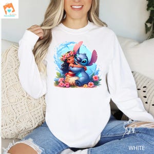 May include: A white long-sleeved shirt with a graphic of Stitch from Lilo & Stitch hugging a girl with a flower lei. The girl is wearing a blue dress and has brown hair. The graphic is set against a blue and white wave background.