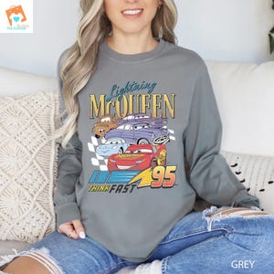 May include: A grey long-sleeved sweatshirt featuring a graphic of Lightning McQueen and other Cars characters. The text "Lightning McQueen", "Think Fast", and "95" are printed on the shirt.