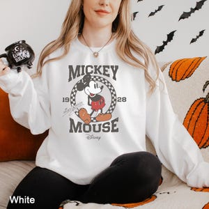 Puede incluir: Sudadera blanca con un gr&aacute;fico de Mickey Mouse. El gr&aacute;fico presenta a Mickey Mouse con sus cl&aacute;sicos pantalones cortos rojos y zapatos amarillos. El texto "Mickey Mouse" est&aacute; impreso en negro sobre el gr&aacute;fico, y el texto "1928" est&aacute; impreso en negro a la derecha del gr&aacute;fico. El texto "Disney" est&aacute; impreso en negro debajo del gr&aacute;fico.