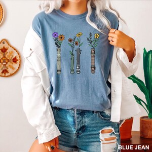 Puede incluir: Una camiseta azul con un gr&aacute;fico de cuatro sables de luz con flores que crecen desde la parte superior. Los sables de luz son de diferentes colores, incluyendo verde, azul, rojo y amarillo.