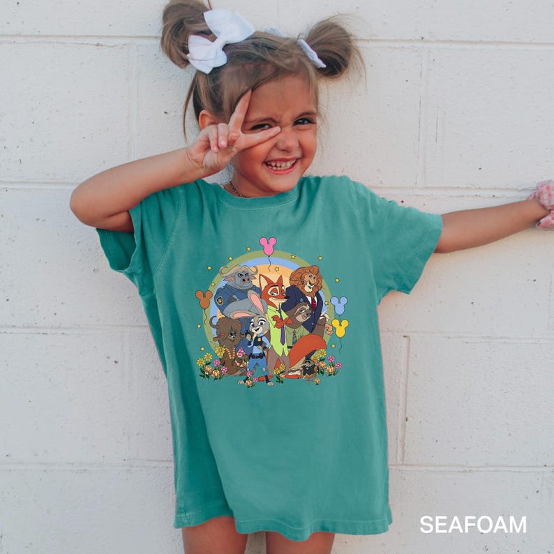 May include: A teal-colored t-shirt featuring a colorful graphic of characters from the animated movie Zootopia. The graphic includes Judy Hopps, Nick Wilde, and other characters. The text "SEAFOAM" is printed at the bottom of the shirt.