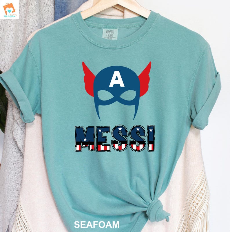 May include: A light blue t-shirt with a red, white, and blue American flag design. The design features a superhero mask with the letter "A" on the forehead and the word "MESSI" below the mask.