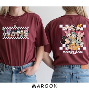 May include: A maroon t-shirt featuring a front graphic of Disney characters in a row over a white and maroon checkerboard pattern, and a back graphic of the same characters clustered over a checkerboard with the text MICKEY & CO. EST 1928.