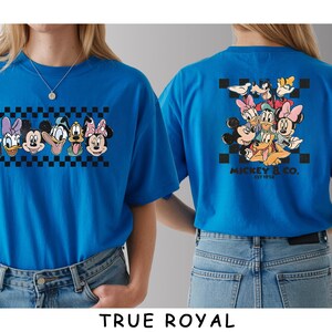 May include: A true royal blue t-shirt featuring a front graphic of six Disney character faces arranged in a horizontal row over a black and white checkerboard pattern. The back displays a larger group of characters with the text MICKEY & CO. EST 1928.