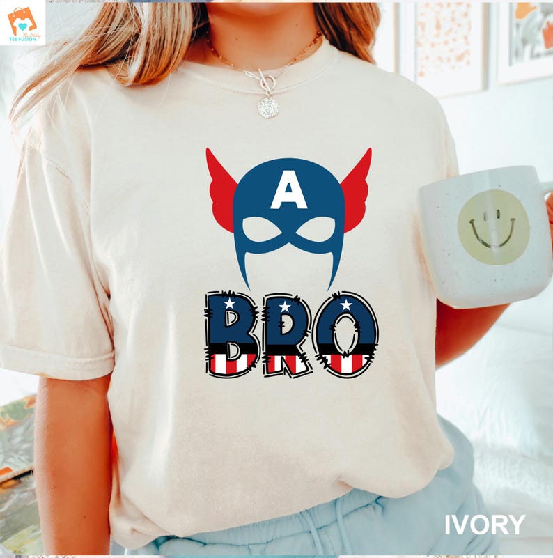 May include: A white t-shirt with a blue and red American flag design. The design features a superhero mask with the letter "A" on the forehead and the word "BRO" below it.