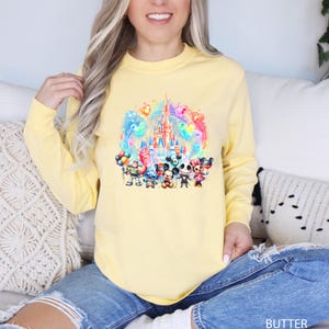 Puede incluir: Una sudadera amarilla de manga larga con un estampado gr&aacute;fico colorido de un castillo de Disney, Mickey Mouse, Minnie Mouse, Stitch y Jack Skellington. Los personajes sostienen globos y est&aacute;n de pie frente al castillo.
