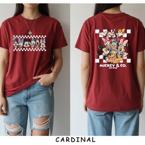 May include: A cardinal red short-sleeve t-shirt featuring a graphic of Disney characters on the front and back. The front shows a row of character faces over a white and red checkerboard pattern. The back displays the same characters with the text MICKEY & CO. EST 1928.