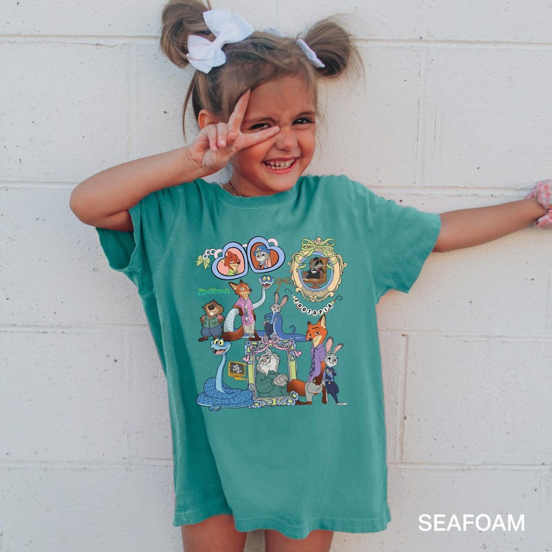 May include: A teal-colored t-shirt featuring a colorful cartoon graphic of characters from Zootopia. The graphic includes images of Nick Wilde, Judy Hopps, and other characters. The shirt is worn by a young girl.
