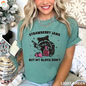 May include: A light green t-shirt with a black and white illustration of a raccoon holding a jar of strawberry jam and a gun. The text on the shirt reads "Strawberry Jams But My Glock Don't".