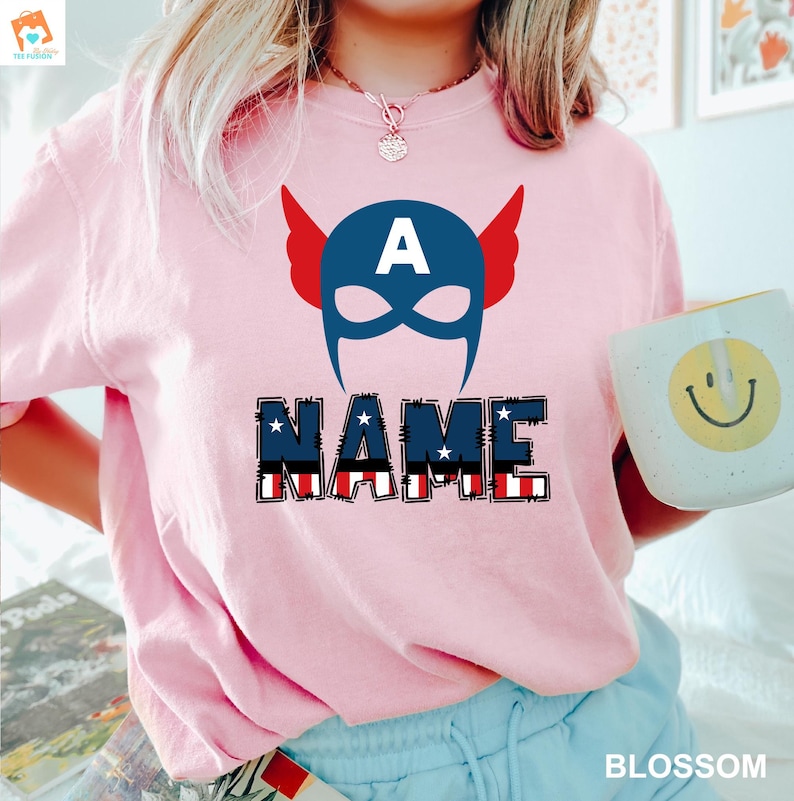 May include: Pink t-shirt with a red, white, and blue American flag design. The shirt features a superhero mask with the letter "A" on the forehead and the word "NAME" in a stylized font.