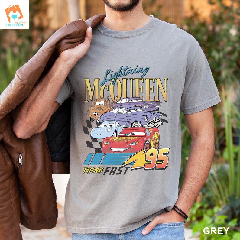 May include: A grey t-shirt featuring a graphic of Lightning McQueen and other Cars characters. The text "Lightning McQueen" is printed in yellow and the text "Think Fast" is printed in black. The number "95" is printed in red.