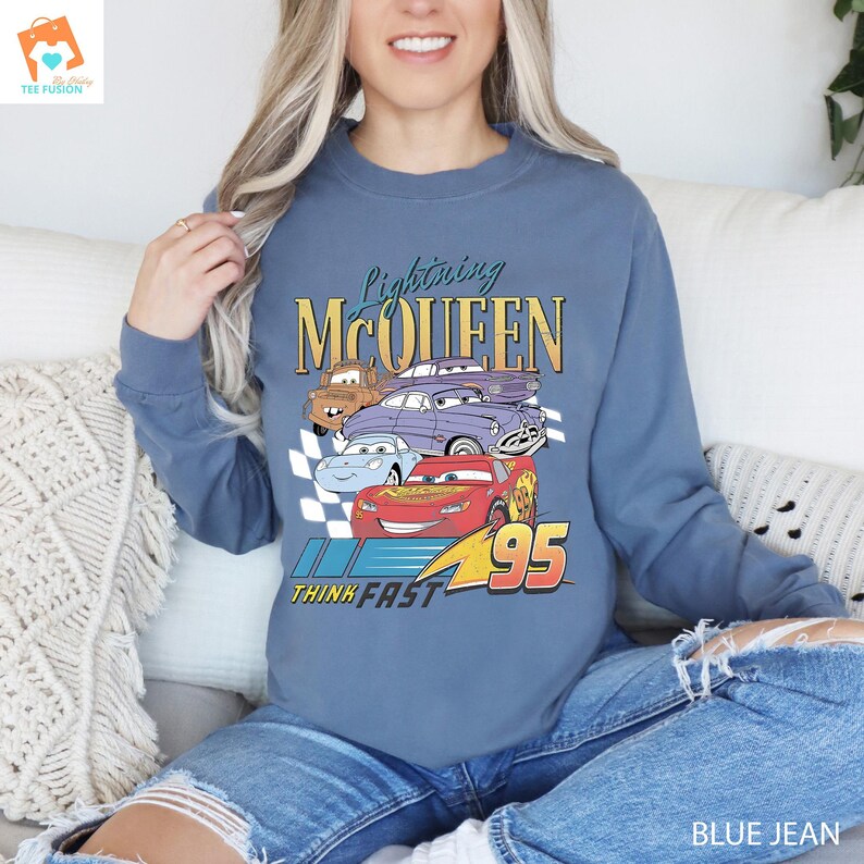 May include: A blue sweatshirt featuring a vintage-style graphic of Lightning McQueen and other Cars characters. The graphic includes the text "Lightning McQueen", "Think Fast", and "95".