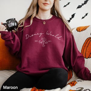 May include: Maroon crewneck sweatshirt with the text "Disney World" in a script font, with a Mickey Mouse graphic and the text "EST. 1971". The sweatshirt is a solid maroon color.