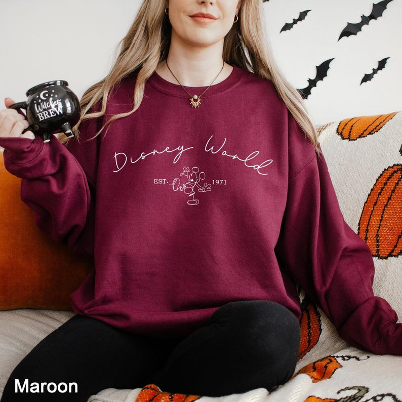 May include: Maroon crewneck sweatshirt with the text "Disney World" in a script font, with a Mickey Mouse graphic and the text "EST. 1971". The sweatshirt is a solid maroon color.