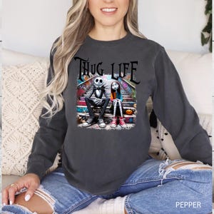 Puede incluir: Una sudadera gris oscuro con un estampado gr&aacute;fico de Jack Skellington y Sally de Pesadilla antes de Navidad sentados en un tramo de escaleras. El texto "THUG LIFE" est&aacute; impreso encima de ellos.
