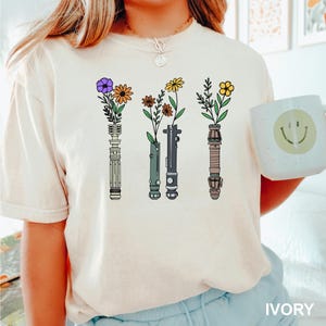 Puede incluir: Una camiseta blanca con un gr&aacute;fico de cuatro sables de luz con flores que crecen desde la parte superior. Los sables de luz son verdes, negros y morados. El texto "IVORY" est&aacute; en la esquina inferior derecha de la imagen.