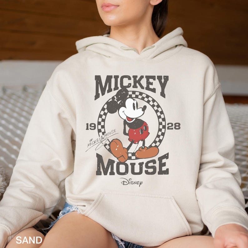 Puede incluir: Una sudadera con capucha de color arena con un gr&aacute;fico vintage de Mickey Mouse. El gr&aacute;fico presenta a Mickey Mouse con sus caracter&iacute;sticos pantalones cortos rojos y guantes blancos, de pie en un c&iacute;rculo a cuadros con el texto "Mickey Mouse" y "1928" encima y "Disney" debajo.