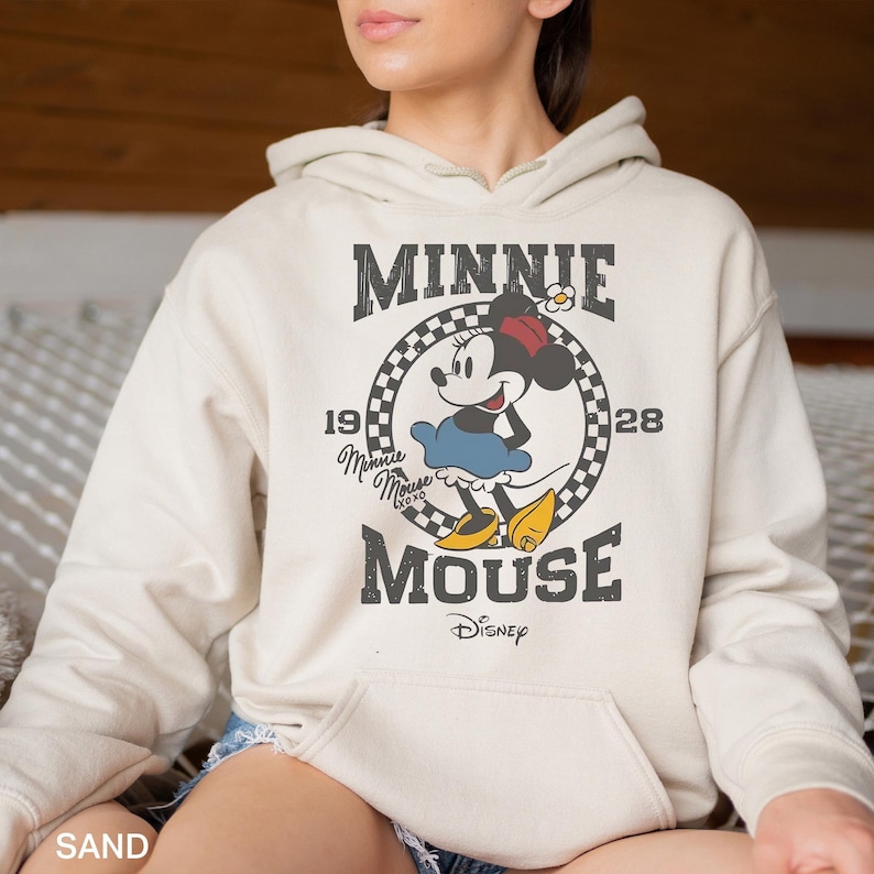 Puede incluir: Una sudadera con capucha de color crema con un gr&aacute;fico de estilo vintage de Minnie Mouse. El gr&aacute;fico presenta a Minnie Mouse con un vestido rojo y zapatos amarillos, con el texto "Minnie Mouse" y "Disney" en negro.