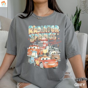 May include: A gray t-shirt with a graphic print of cartoon cars from the movie Cars. The text "Radiator Springs" is printed in a vintage style font.