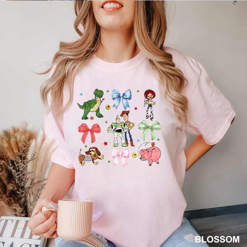 May include: A pink t-shirt with a graphic print of characters from the Toy Story movie. The characters include Woody, Buzz Lightyear, Rex, Hamm, and Jessie. The graphic also includes several colorful bows.