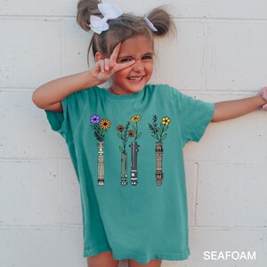 Puede incluir: Una camiseta azul turquesa con un gr&aacute;fico de tres sables de luz con flores que crecen de ellos. El texto "SEAFOM" est&aacute; en la esquina inferior derecha de la imagen.