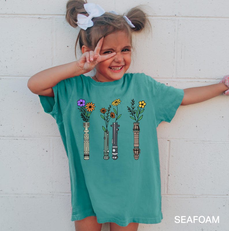 Puede incluir: Una camiseta azul turquesa con un gr&aacute;fico de tres sables de luz con flores que crecen de ellos. El texto "SEAFOM" est&aacute; en la esquina inferior derecha de la imagen.