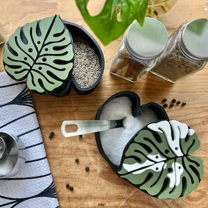 May include: Two green and white ceramic leaf-shaped containers with lids. One container is filled with white salt and the other with black peppercorns. The containers are on a wooden surface with a white and black patterned kitchen towel, two measuring spoons, and a small pile of black peppercorns.