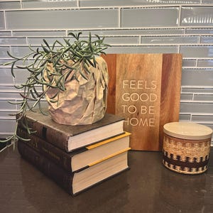 May include: A stack of three books with a brown paper bag planter with a green succulent plant on top. The books have yellow edges. A wooden sign with the text "Feels good to be home" is on the right. A brown and white woven candle jar is on the counter.