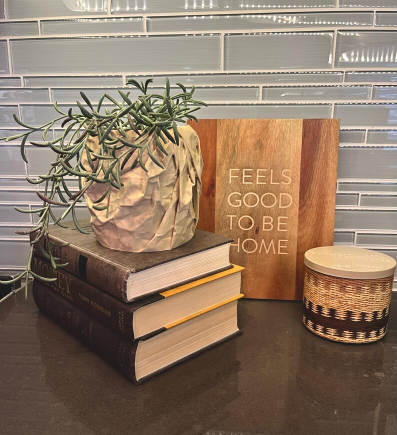 May include: A stack of three books with a brown paper bag planter with a green succulent plant on top. The books have yellow edges. A wooden sign with the text "Feels good to be home" is on the right. A brown and white woven candle jar is on the counter.