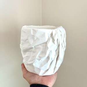 May include: White ceramic planter with a crumpled, textured surface. The planter is held in a hand.