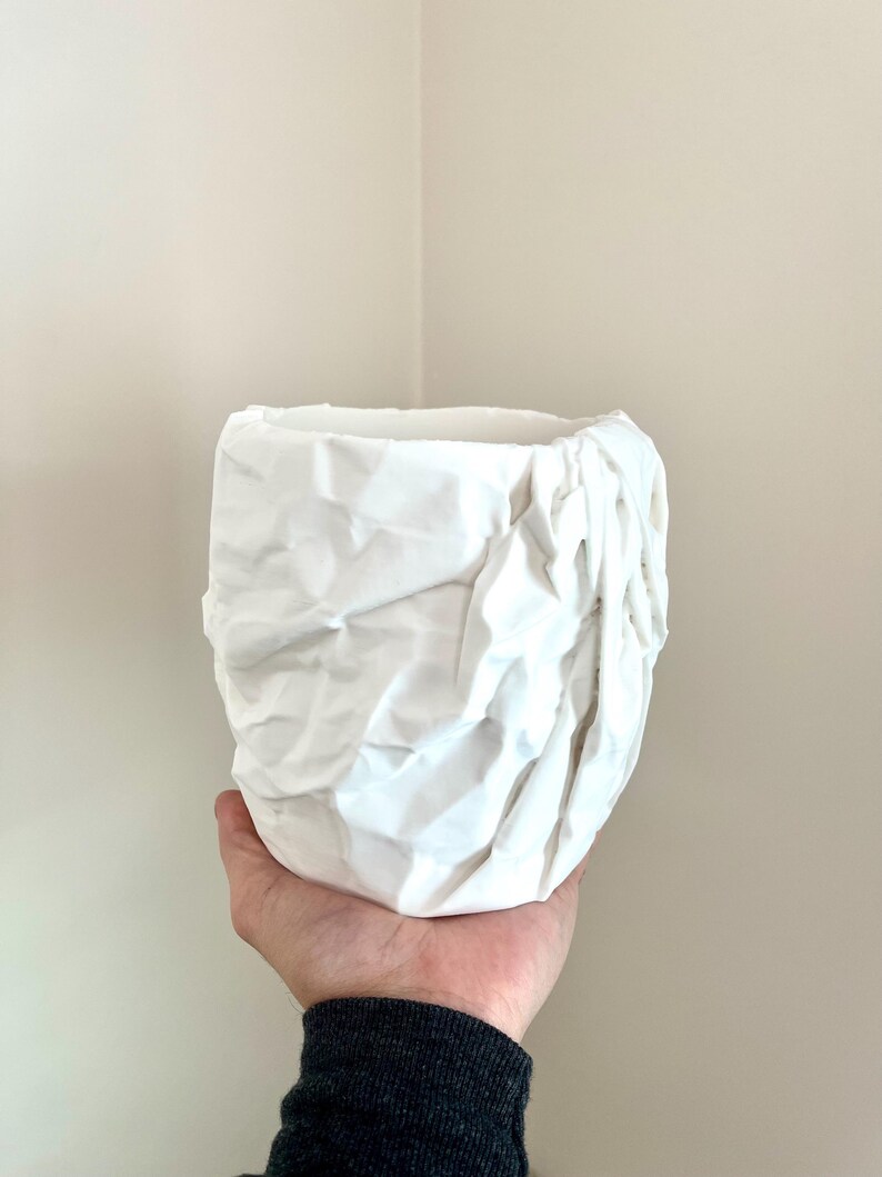 May include: White ceramic planter with a crumpled, textured surface. The planter is held in a hand.