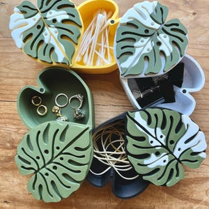 May include: Six small, open, heart-shaped boxes with a monstera leaf design. The boxes are painted in different colors, including green, yellow, black, and white. The boxes are filled with various items, such as cotton swabs, hair ties, and jewelry.