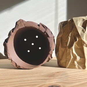 May include: Two ceramic planters, one brown and one beige, with a crumpled paper texture. Both planters have drainage holes.