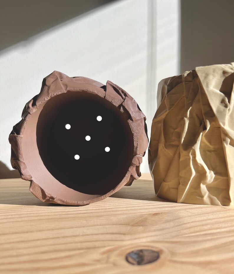 May include: Two ceramic planters, one brown and one beige, with a crumpled paper texture. Both planters have drainage holes.