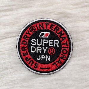 May include: Circular embroidered patch with a black central design. The patch features the words "SUPER DRY" and "JPN" in white. The outer ring is red with "SUPERDRY INTERNATIONAL" in white. A white border surrounds the design.