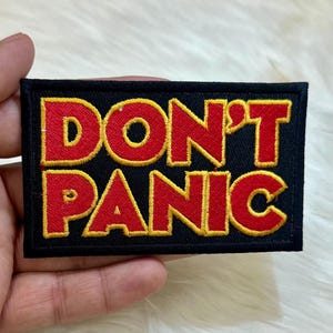 May include: A black fabric patch with yellow stitching around the edges. The patch has the words "DON'T PANIC" in red letters with a yellow outline.