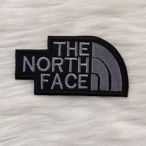 May include: A black and grey embroidered patch with the words "THE NORTH FACE." The patch is rectangular with a curved right edge. The text is grey, and the background is black.