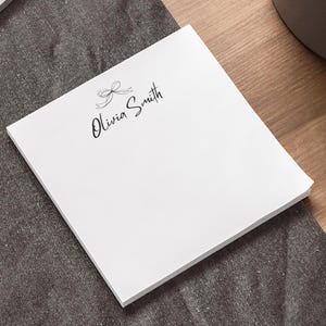 May include: A white notepad with a bow and the name "Olivia Smith" printed in black script. The notepad is on a dark gray surface next to a laptop and a pen. A small potted plant is in the background.