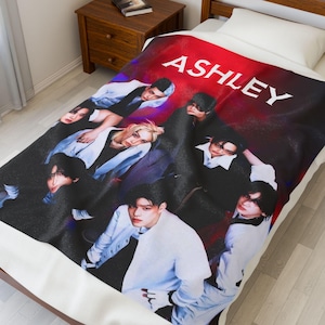 May include: A blanket featuring a group of people in various poses, with the name "ASHLEY" in large white letters against a red background. The blanket is on a bed with a white sheet.