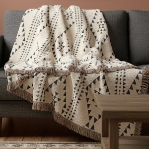 May include: A cream-colored throw blanket with a geometric triangle pattern in black, draped over a gray sofa. The blanket has a fringed edge in a brown hue, adding a decorative touch to the home decor.