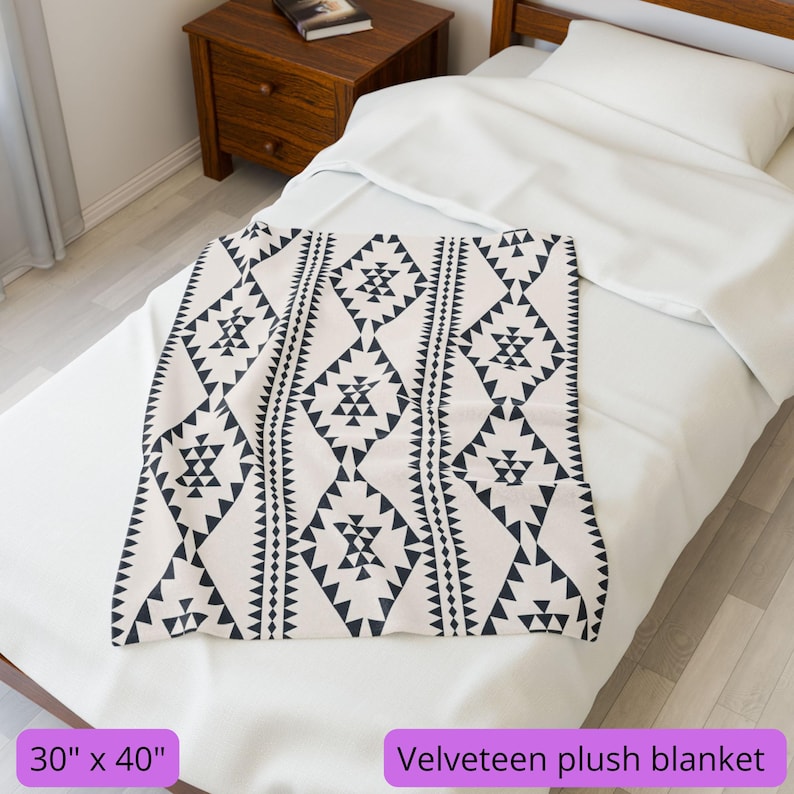 Puede incluir: Una manta de felpa de terciopelo crema con un patr&oacute;n geom&eacute;trico negro, de 76 x 102 cm. La manta est&aacute; doblada sobre una cama blanca, con el texto "Velveeteen plush blanket" visible.