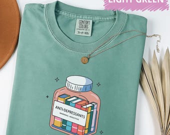 May include: Light green t-shirt with a graphic of a pill bottle filled with books labeled "ANTI-DEPRESSANTS" and "WARNING: ADDICTIVE." The text "School psych" is below the graphic. A silver necklace with a small pendant is draped across the shirt.