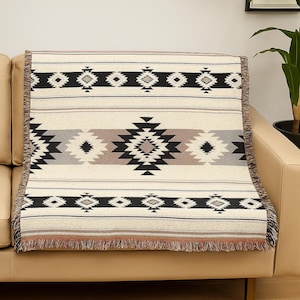 Op de afbeelding: Crèmekleurige geweven sprei met een geometrisch patroon in zwart, grijs en bruin. De deken heeft een centraal ruitontwerp en horizontale strepen, met gefranjerde randen. De deken is over een beige bank gedrapeerd.