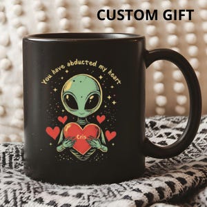 May include: Black coffee mug with a graphic of a green alien holding a red heart with the text "You have abducted my heart" and the name "Cris" on the heart.
