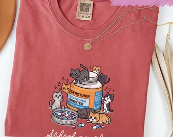 May include: Crimson-colored t-shirt with a graphic of cartoon cats surrounding a bottle labeled "Serotonin Booster." The shirt also features the text "School psych."