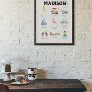 May include: A framed print with the word "MADISON" in bold black letters at the top. The print features illustrations of various landmarks and attractions in Madison, Wisconsin, including the State Capitol, the Farmers Market, the Memorial Union, the Bigfoot  Hangover, the  Alliant Energy Center, the  Local Craft Beer, the  State Street, and the  Jump Around. The print also includes a small outline of the state of Wisconsin.