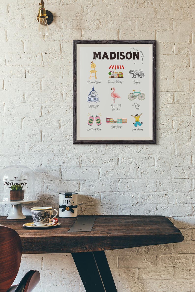 May include: A framed print with the word "MADISON" in bold black letters at the top. The print features illustrations of various landmarks and attractions in Madison, Wisconsin, including the State Capitol, the Farmers Market, the Memorial Union, the Bigfoot  Hangover, the  Alliant Energy Center, the  Local Craft Beer, the  State Street, and the  Jump Around. The print also includes a small outline of the state of Wisconsin.