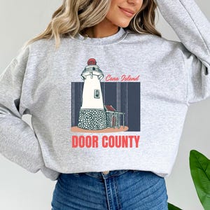 Puede incluir: Sudadera gris con un gráfico de un faro en la isla de Cana en el condado de Door, Wisconsin. El faro es blanco con un techo rojo y está rodeado de rocas grises. El texto "Cana Island" y "Door County" está impreso en rojo.